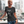 A woman wearing a black graphic t-shirt with the text 'Reach Out, Touch Faith! MATTHEW 6:33, stands on a balcony with a blurred city skyline background.