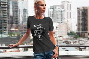 A woman wearing a black graphic t-shirt with the text 'Reach Out, Touch Faith! MATTHEW 6:33, stands on a balcony with a blurred city skyline background.
