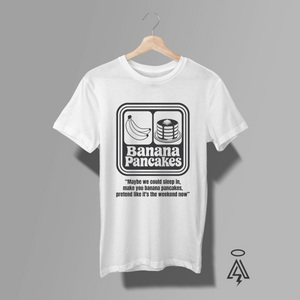 A white short-sleeve t-shirt. The shirt features a black retro-style graphic on the chest with a banana and pancakes illustration, the text 'Banana Pancakes', and a quote-lyric underneath.