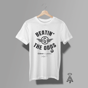 A white short-sleeve t-shirt hanging on a wooden hanger against a solid gray background. The shirt features a black graphic print on the front with the text 'BEATIN' THE ODDS' and a winged 'G' logo, with 'Gideon / (gideon)' and 'judges 7:7' printed below.