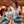 A woman and a girl sitting together in front of a wooden wall with large lit marquee letters. Both are wearing white graphic T-shirts and ripped blue jeans.