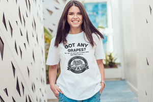 A smiling young woman stands in a bright hallway wearing a white graphic T-shirt with the text 'GOT ANY GRAPES?' and a circular vintage-style emblem on the front, paired with blue jeans.