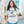 A smiling young woman stands in a modern outdoor walkway wearing a white graphic t-shirt with an angel illustration and the text 'WHAT WOULD BABY JESUS DO?'.